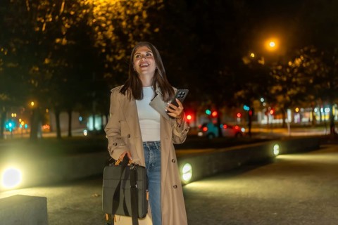 Woman in trench coat walking through an illuminated city park at night, smiling and confident while