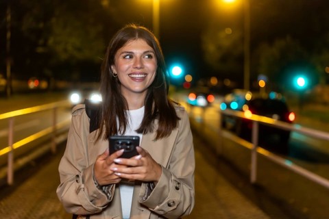 Young woman smiling while holding a smartphone and walking through a downtown street at night,
