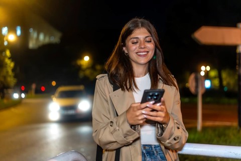 Young brunette woman standing on a city street at night, smiling and checking her smartphone in a