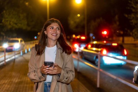 Young woman in a trench coat smiling, holding a phone, and looking away while walking on a city