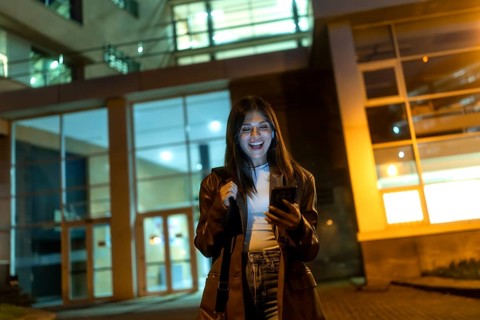Young woman standing on an urban street at night, happily looking at her smartphone while holding a
