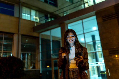 Young woman standing on a city street at night, looking at her smartphone and smiling with eyes