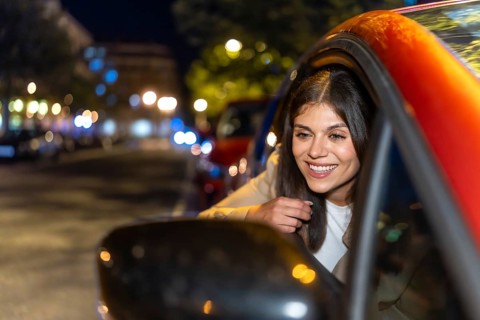Young woman leaning out of a car window at night, smiling as city lights and bokeh blur by,