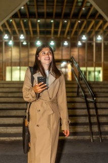 Young woman dressed in a trench coat smiling, holding a mobile phone, and walking up illuminated