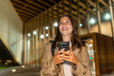 Young woman smiling, holding phone while walking illuminated city streets at night, confidently