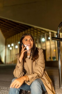 Young woman sitting outdoors at night, smiling brightly while having a phone conversation,