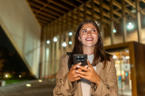 Young woman smiling while holding a smartphone and looking up, exploring a modern city street at