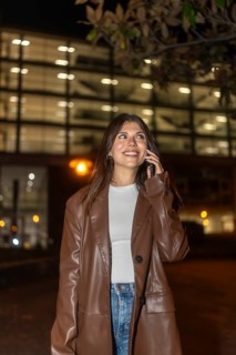 Young woman in a brown leather jacket smiling, talking on a mobile phone in an urban street at