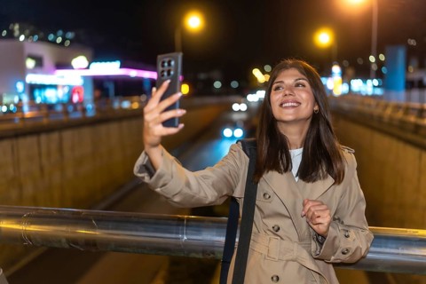 Young woman smiling and holding a smartphone, capturing a selfie against the bright, blurred lights