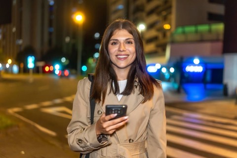 Woman smiling brightly and holding a smartphone, standing on a bustling city street at night with