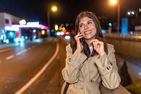 Young woman in a trench coat smiling as she talks on her mobile phone on a bustling city street at