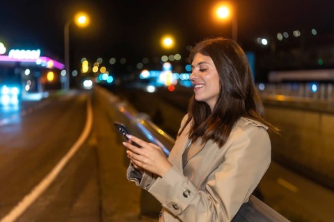 Young woman standing on an urban bridge at night, smiling while interacting with her smartphone,