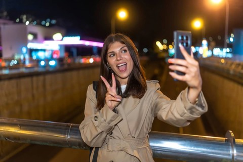 Young woman wearing a trench coat standing on a bridge, holding a smartphone and taking a self