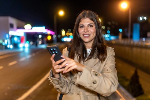 Young woman in a trench coat standing on a city street at night, smiling at her smartphone as she