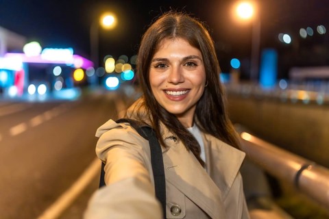 Young woman smiling and holding a smartphone, capturing a selfie portrait outdoors in an urban