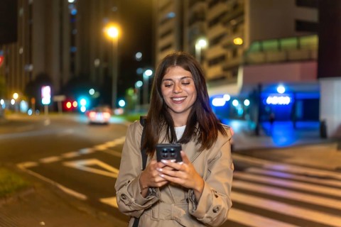 Young woman smiling with eyes closed, enjoying her smartphone on a city street at night, relaxed