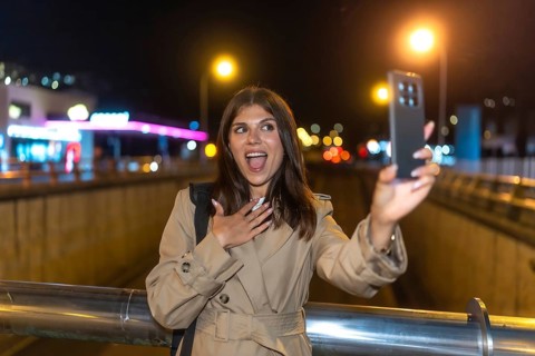 Young woman in trench coat surprised and joyful taking a nighttime selfie on a bustling city
