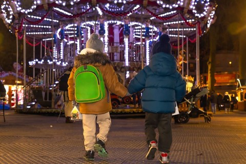 Two young brothers, holding hands, walk away from a brightly lit carousel at a christmas market in