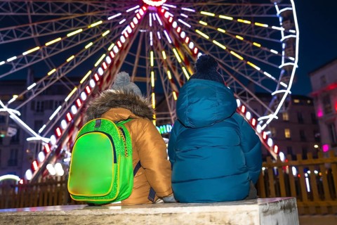 Two children with winter coats and backpacks admiring a brightly lit ferris wheel at a christmas