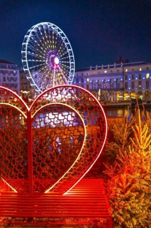 Illuminated heart shaped bench with small christmas trees and ferris wheel in the background,