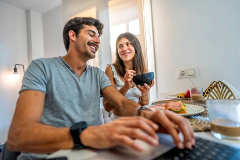 Young couple enjoying breakfast in a bright kitchen, working from home on a laptop while sharing