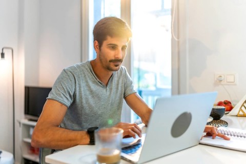 Young man working from home, typing on a laptop and jotting down notes while basking in the warm