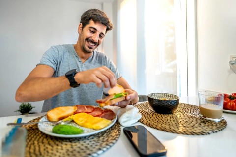 Young man smiling while preparing a delicious sandwich for breakfast at home, enjoying a healthy