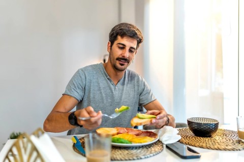 Man adding fresh avocado to toast while enjoying a healthy breakfast at home, embracing a balanced