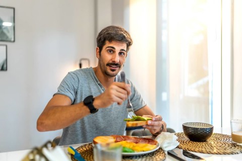 Happy man enjoying avocado toast alongside ham and eggs while relaxing in his modern apartment,