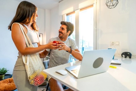Young woman arriving home from grocery shopping, giving an apple to her boyfriend working remotely