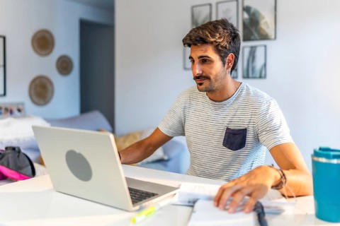Young male freelancer working remotely at home, using a laptop and reviewing notes while sitting