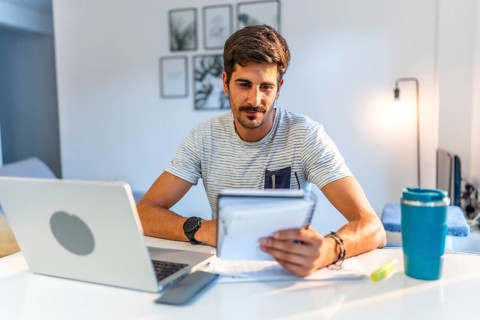Young man managing his business remotely, reviewing notes and working on his laptop in his bright