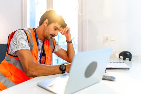 Young construction worker feeling stressed while working remotely from home using laptop, having