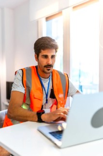 Focused engineer wearing high visibility vest working on laptop in bright modern office, managing