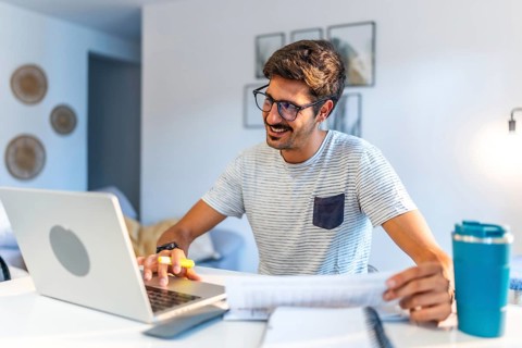 Young man working comfortably at home, engaging with a laptop and reviewing documents, smiling