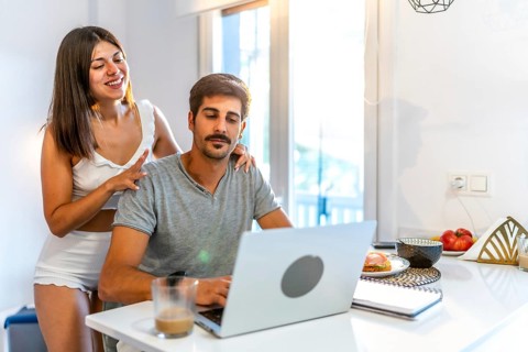 Young woman massaging her husband's shoulders while he's working from home on his laptop in the