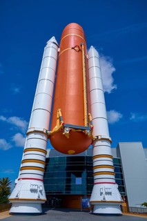 Large orange space shuttle with booster rockets in front of a modern building, Kennedy Space Center