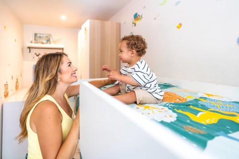Smiling mother looking at her cute baby sitting on a bunk bed in a decorated bedroom, happy family