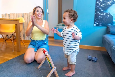 Mother encouraging her baby boy to walk, clapping hands in living room, promoting child development