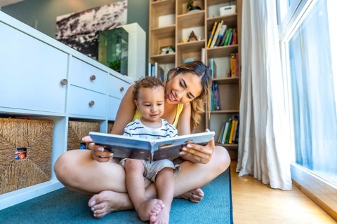 Happy mother spending quality time reading a book to her baby son, sitting on the floor in the
