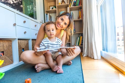 Happy mother and her baby son reading a book together, sitting on the floor in the living room at