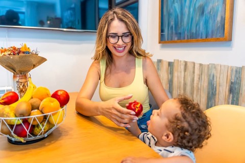 Loving mother giving a fresh peach to her baby son while sitting at the kitchen table, promoting