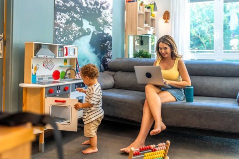 Woman working remotely on laptop while her toddler plays with a toy kitchen, demonstrating work