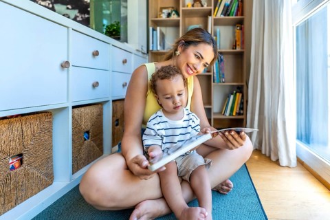Happy mother and baby son reading a book together, sitting on the floor in the living room at home