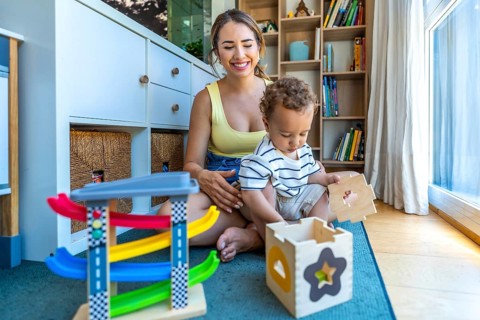 Smiling mother watching her cute toddler son playing with a shape sorter toy and a colorful car