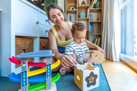 Happy mother and son are sitting on the floor playing with a wooden shape sorter and colorful toy
