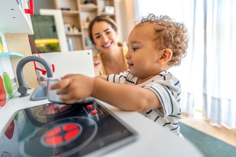 Happy toddler playing with toy kitchen while his mother watches him, promoting early childhood