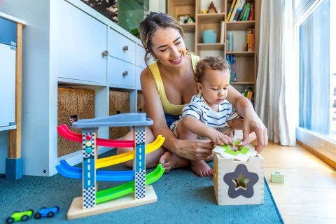 Happy mother helping her baby son playing with a shape sorter toy, sitting on a play mat in a
