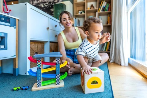 Happy mother watching her toddler son playing with educational wooden toys in a modern living room