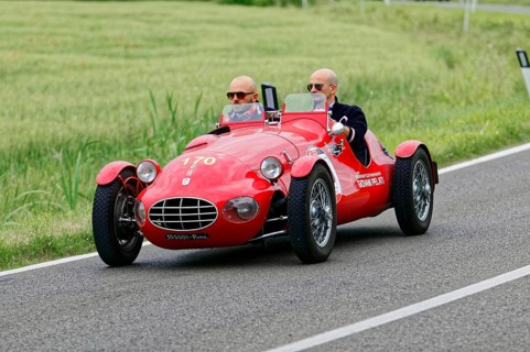 An open red sports car with two drivers drives through a green landscape, classic car, car race,